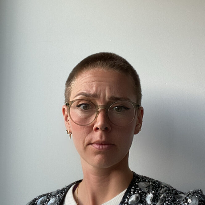 White woman with a bald head and glasses wearing a silver jacket looking straight into the camera
